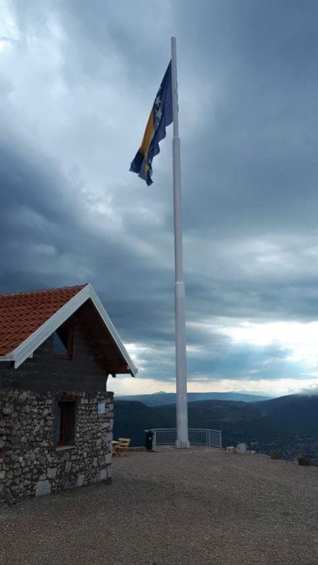 Zastava Bosne i Hercegovine na visokom jarbolu na Fortici iznad Mostara, uz kamenu kuću i pogled na dolinu